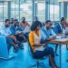 A cinematic editorial photo of a diverse group of young Kenyan tech professionals collaborating energetically around laptops and digital devices in a modern, sunlit Nairobi coworking space, symbolizing innovation and top IT talent in Kenya’s booming tech staffing industry.